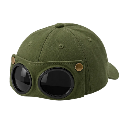 An olive green baseball cap styled with a vintage aviator helmet front, featuring two large round black goggles with black rims and brass-tone rivets on the side, shown on a transparent checkered background.